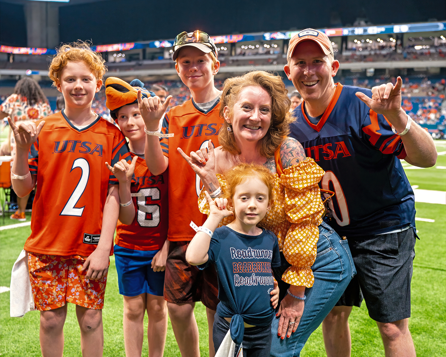 A family holding the schools "birds up" hand gesture and smiling for a photo, From left to right, Four children, a mother, and a father, huddled together posing.